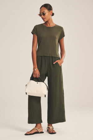 A woman wears the CASEY TEE by Velvet by Graham & Spencer, an olive green linen knit top, styled with wide-leg pants, sandals, sunglasses, and a white handbag.
