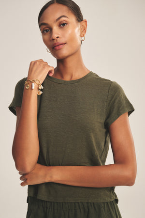 A woman in the CASEY TEE by Velvet by Graham & Spencer, an olive green linen knit with a high-low hem, stands with one arm raised to show off her gold charm bracelet.