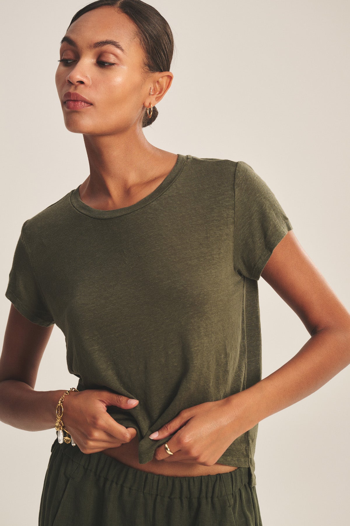 A woman wearing the olive green CASEY TEE by Velvet by Graham & Spencer with matching pants stands against a neutral background, holding the hem of her shirt with one hand.-41338477740225