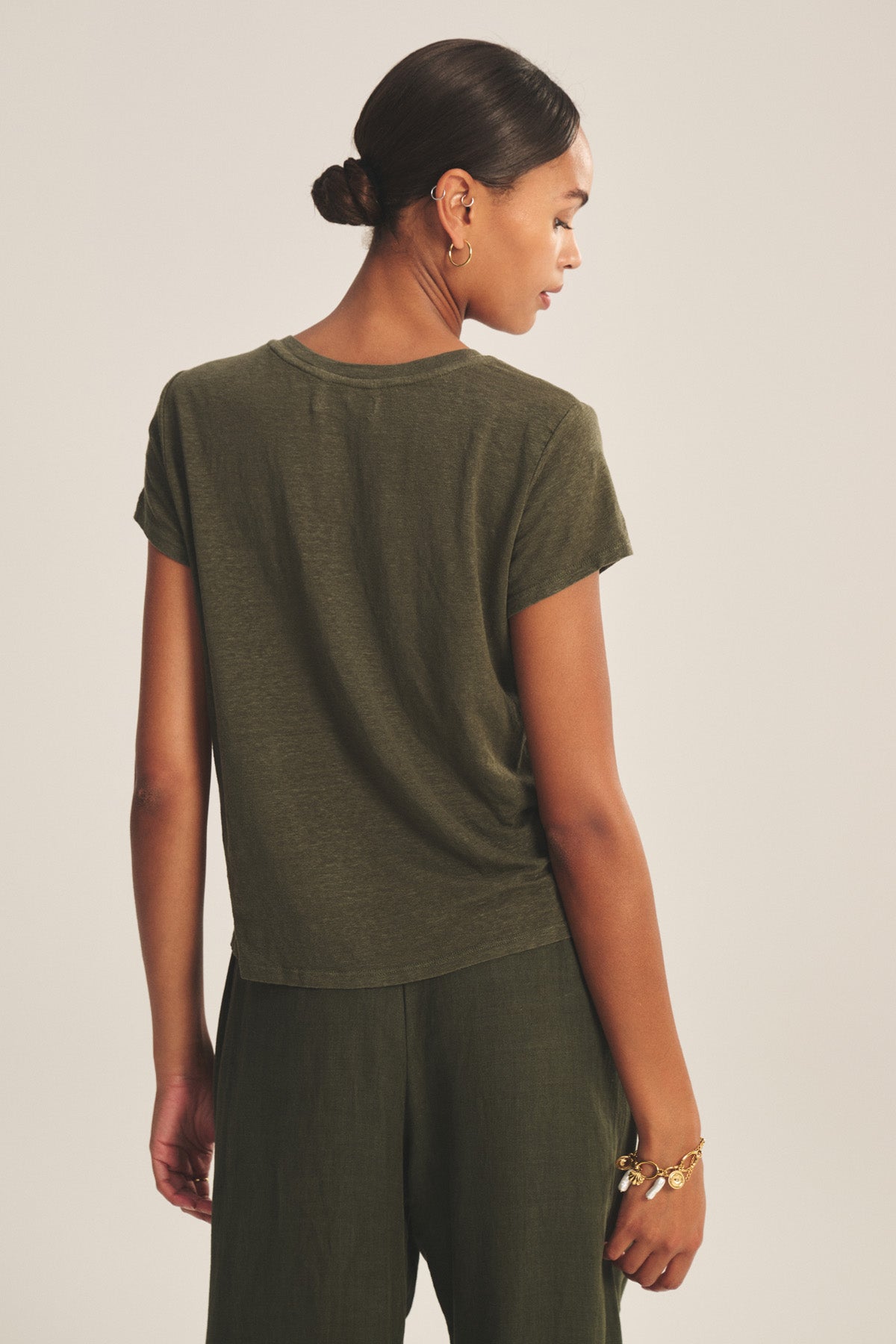 A woman with dark hair in a low bun wears the Velvet by Graham & Spencer CASEY TEE in olive green linen knit, paired with matching pants, and stands with her back to the camera.-41338477674689