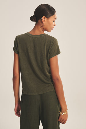 A woman with dark hair in a low bun wears the Velvet by Graham & Spencer CASEY TEE in olive green linen knit, paired with matching pants, and stands with her back to the camera.