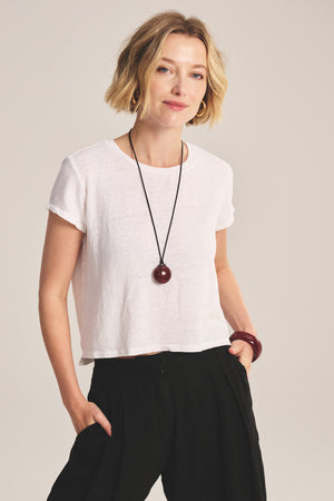 A woman with short blonde hair wears the CASEY TEE by Velvet by Graham & Spencer, styled with black pants, a statement necklace, and bracelet, standing with one hand in her pocket against a simple background.