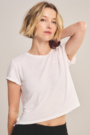 A woman with short blonde hair models the CASEY TEE by Velvet by Graham & Spencer with black pants, posing with one arm raised and hand touching the back of her neck against a plain background.