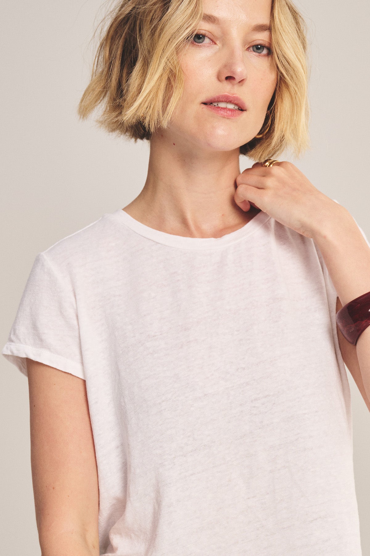A woman with short blonde hair wears the Velvet by Graham & Spencer CASEY TEE, featuring a classic high-low hemline, looking at the camera with a neutral expression and touching her neck.