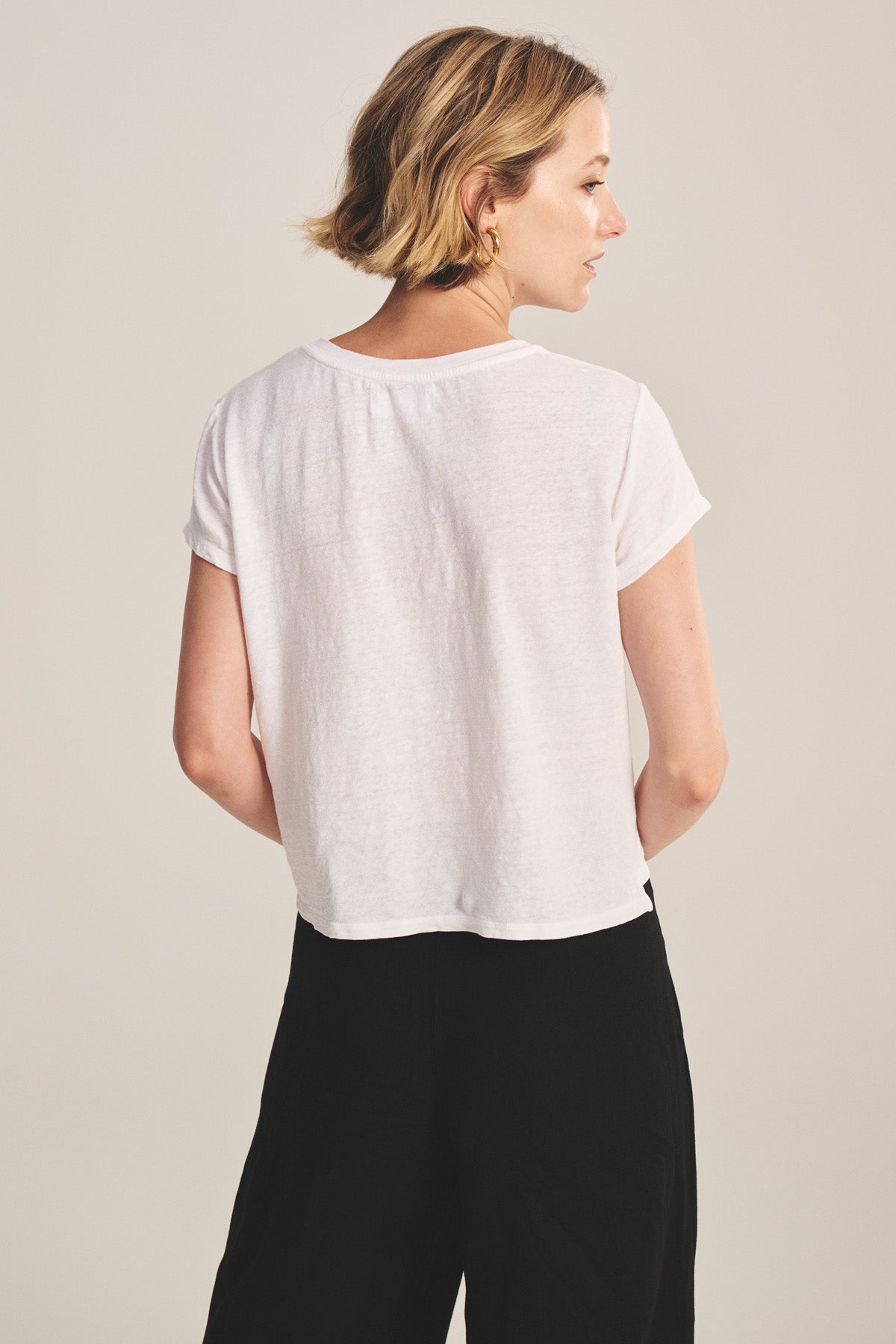 A woman with short blonde hair is seen from behind, wearing the Velvet by Graham & Spencer CASEY TEE with a high-low hemline and black pants against a plain background.