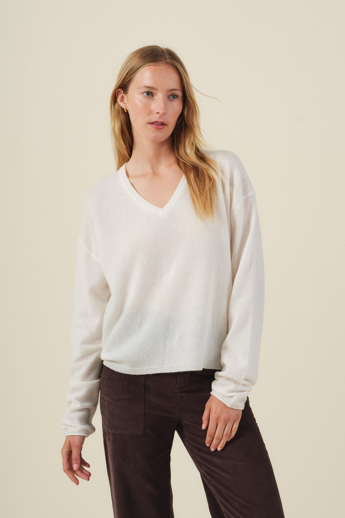 A woman stands against a plain, light-colored background wearing the AMIKA CASHMERE SWEATER by Velvet by Graham & Spencer—an elegant white cashmere sweater with a deep V neckline—paired with dark pants.