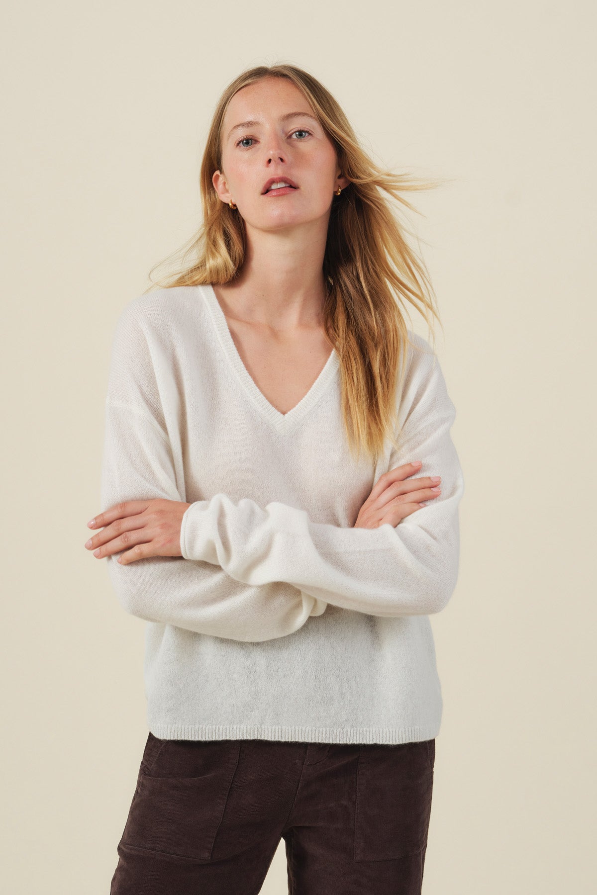 A woman with long blonde hair, standing with arms crossed against a plain light background, wears the AMIKA CASHMERE SWEATER by Velvet by Graham & Spencer—a soft white cashmere knit featuring a V neckline—paired with dark pants.