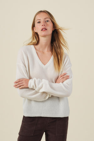 A woman with long blonde hair, standing with arms crossed against a plain light background, wears the AMIKA CASHMERE SWEATER by Velvet by Graham & Spencer—a soft white cashmere knit featuring a V neckline—paired with dark pants.