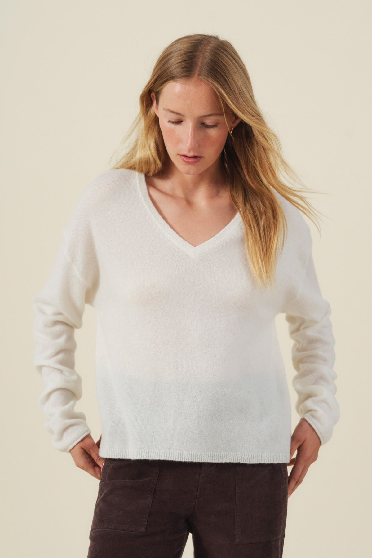 A woman with long blonde hair wears the AMIKA CASHMERE SWEATER by Velvet by Graham & Spencer—a soft white knit with a V neckline and an elegant cropped waist—paired with dark pants. She stands against a light background, looking down as her hands rest near her hips, highlighting the sophisticated silhouette of the sweater.-42204966518977