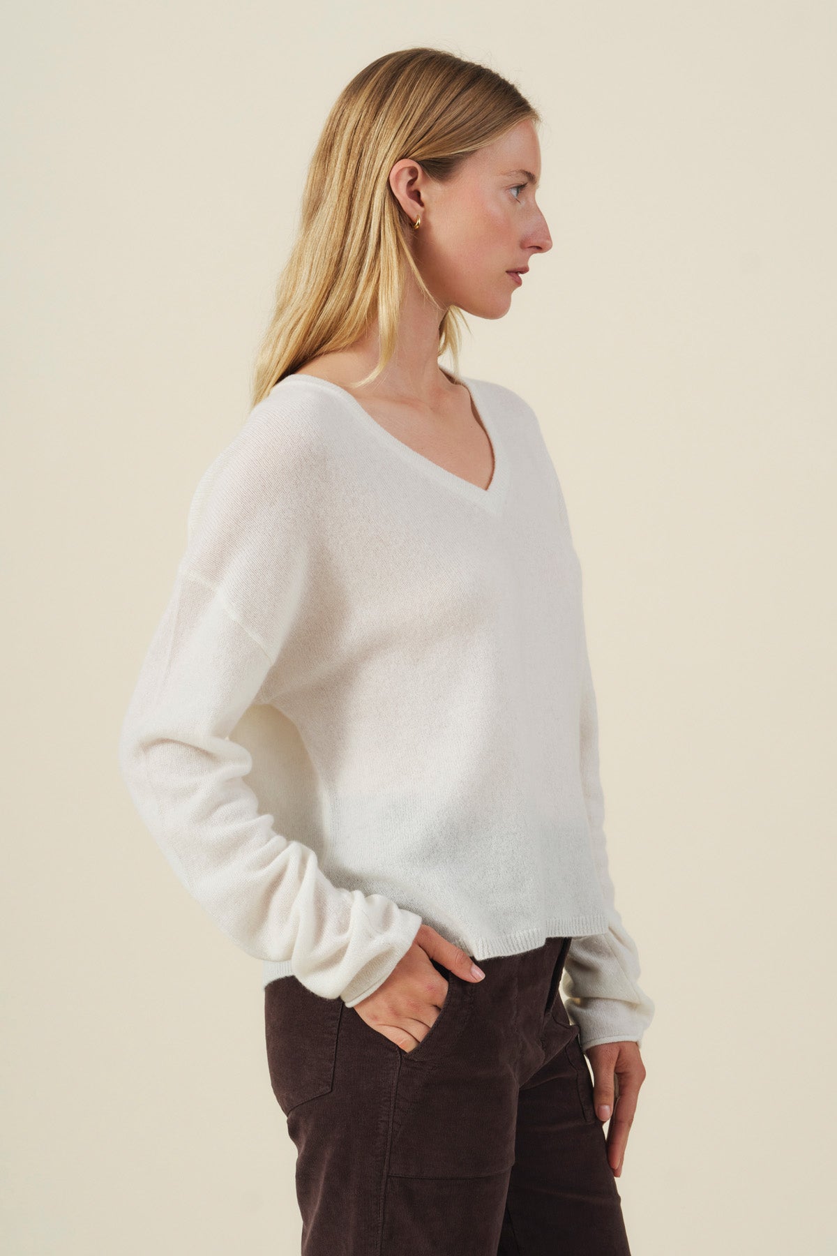 A woman in profile wears the AMIKA CASHMERE SWEATER by Velvet by Graham & Spencer—a white cashmere sweater featuring a deep V neckline—paired with dark pants, standing against a plain beige background with her hands in her pockets.