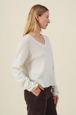 A woman in profile wears the AMIKA CASHMERE SWEATER by Velvet by Graham & Spencer—a white cashmere sweater featuring a deep V neckline—paired with dark pants, standing against a plain beige background with her hands in her pockets.