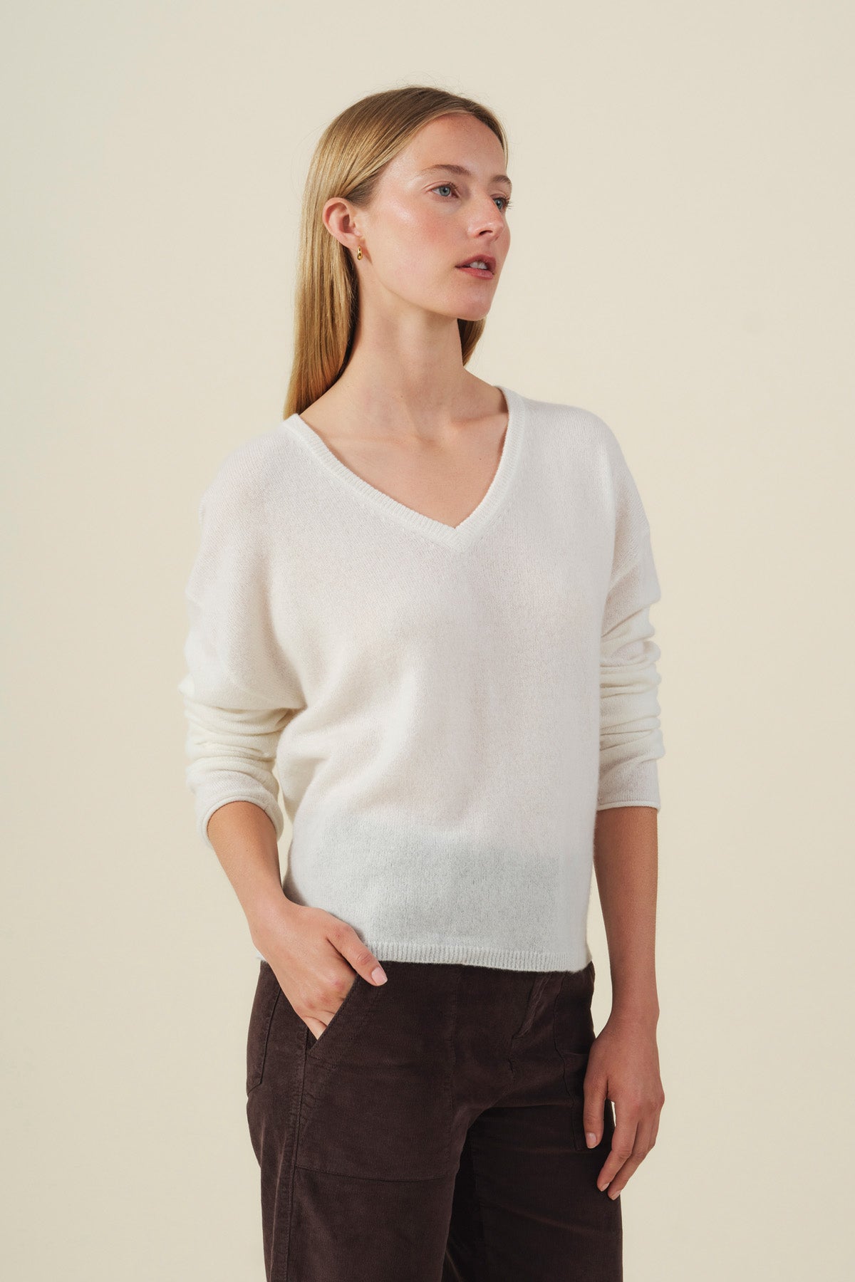 A woman stands against a plain background, wearing the AMIKA CASHMERE SWEATER from Velvet by Graham & Spencer—a white V-neckline sweater crafted from soft cashmere knits—paired with dark pants, her hands in her pockets as she gazes slightly to the side.