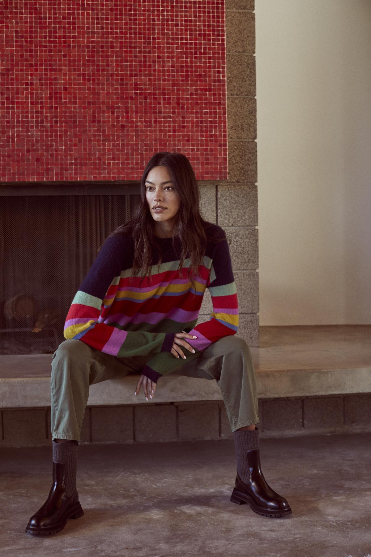 A woman in a colorful sweater sits in front of a fireplace, wearing Velvet by Graham & Spencer's BRYLIE SANDED TWILL UTILITY PANT with patch pockets made of cotton twill.-36118556901569