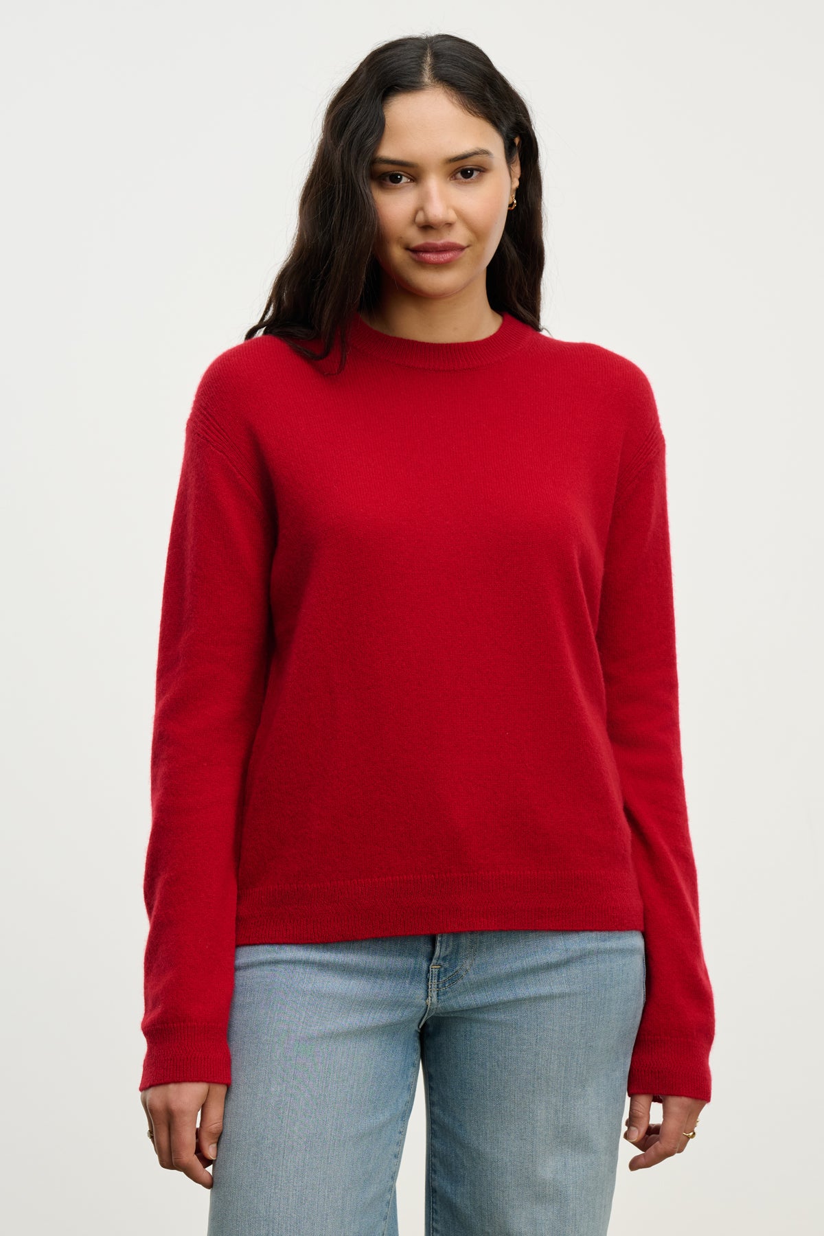   A person with long dark hair wears the Velvet by Graham & Spencer CRYSTAL CASHMERE SWEATER in red and light blue jeans, standing against a plain light background. 
