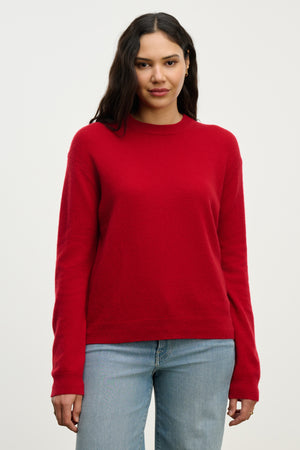 A person with long dark hair wears the Velvet by Graham & Spencer CRYSTAL CASHMERE SWEATER in red and light blue jeans, standing against a plain light background.