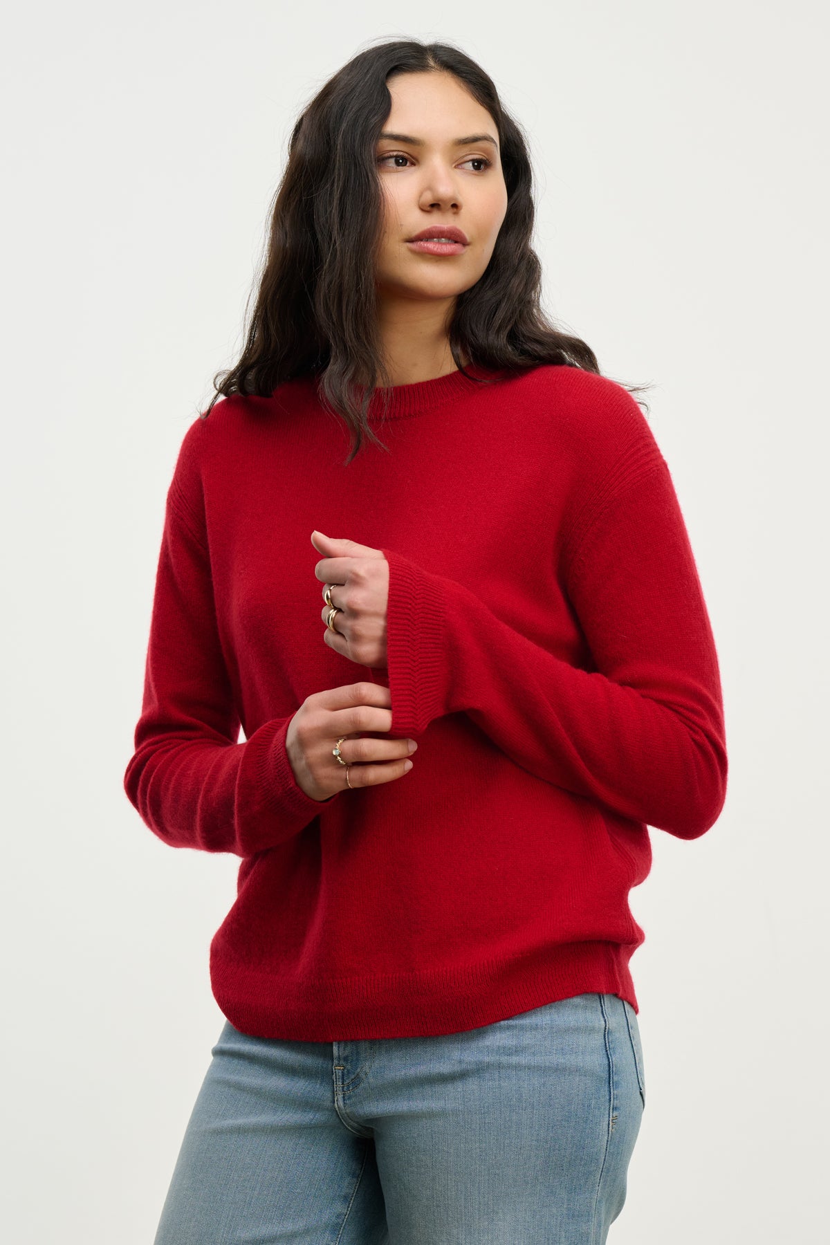   A woman wears the CRYSTAL CASHMERE SWEATER by Velvet by Graham & Spencer—a red crewneck with ribbed trims—paired with light blue jeans, standing against a plain background, looking slightly to the side with hands near her chest. 