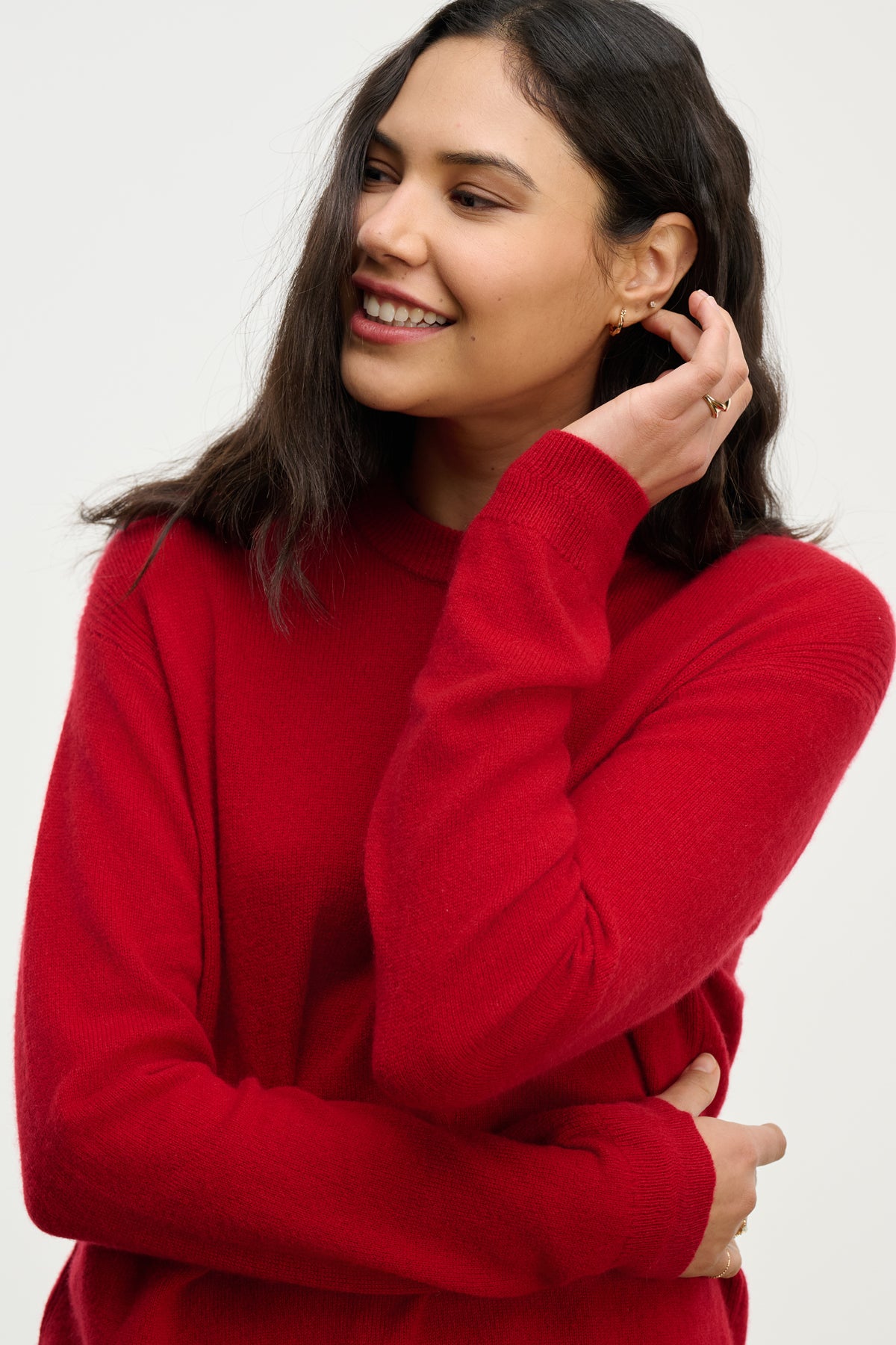   Smiling woman with long dark hair wears the Velvet by Graham & Spencer CRYSTAL CASHMERE SWEATER—red, with a crew neckline and ribbed trims—gazing to the side, one hand touching her ear and the other crossing her body. 