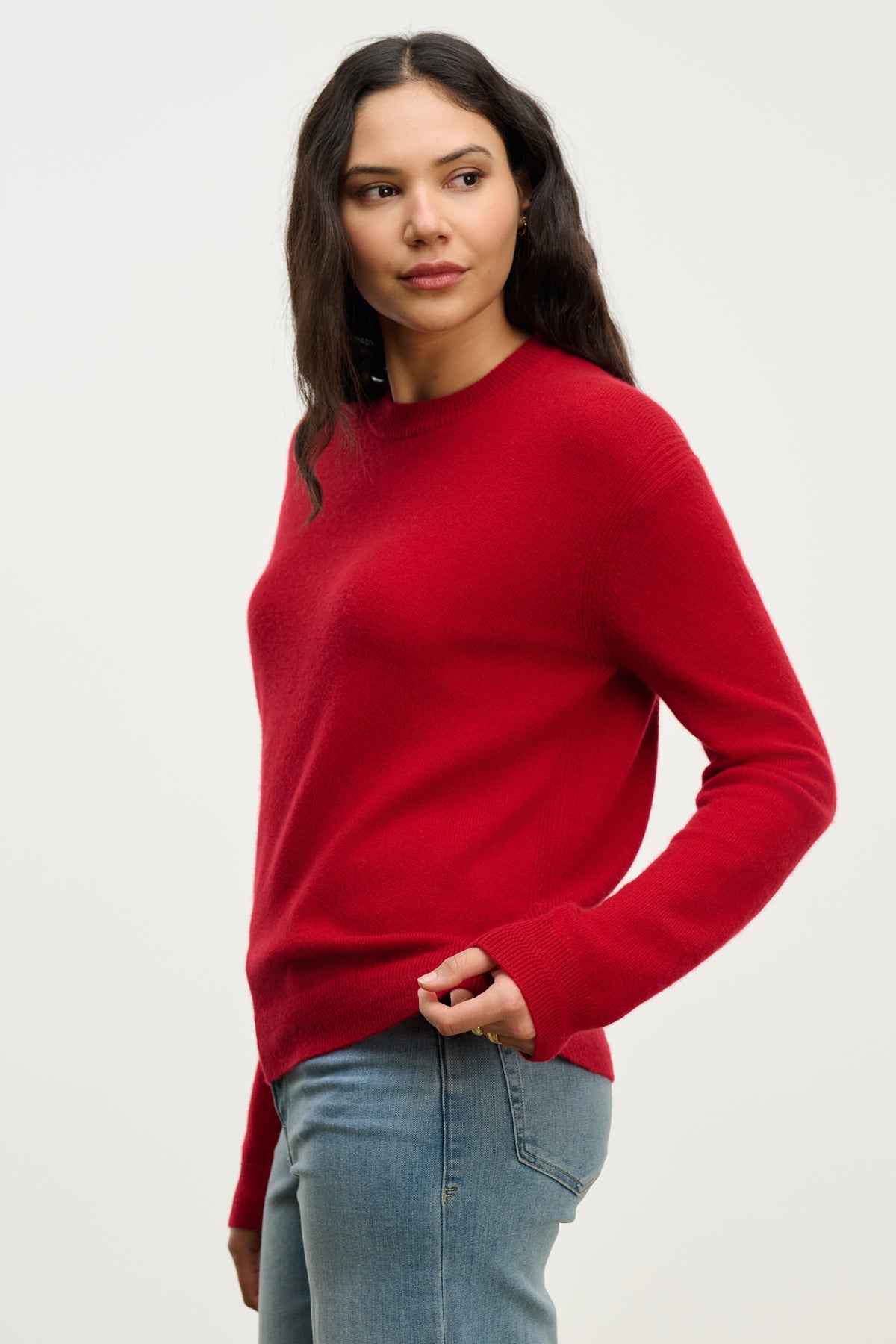   A woman wears the CRYSTAL CASHMERE SWEATER by Velvet by Graham & Spencer, featuring a red hue, crew neckline, and ribbed trims, styled with blue jeans against a plain light background while gazing slightly to the side. 
