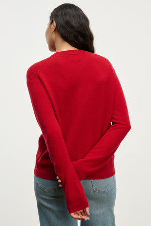 A person with long dark hair wears the Velvet by Graham & Spencer CRYSTAL CASHMERE SWEATER in red, featuring ribbed trims and a crew neckline, styled with blue jeans and standing back to the camera with hands clasped.