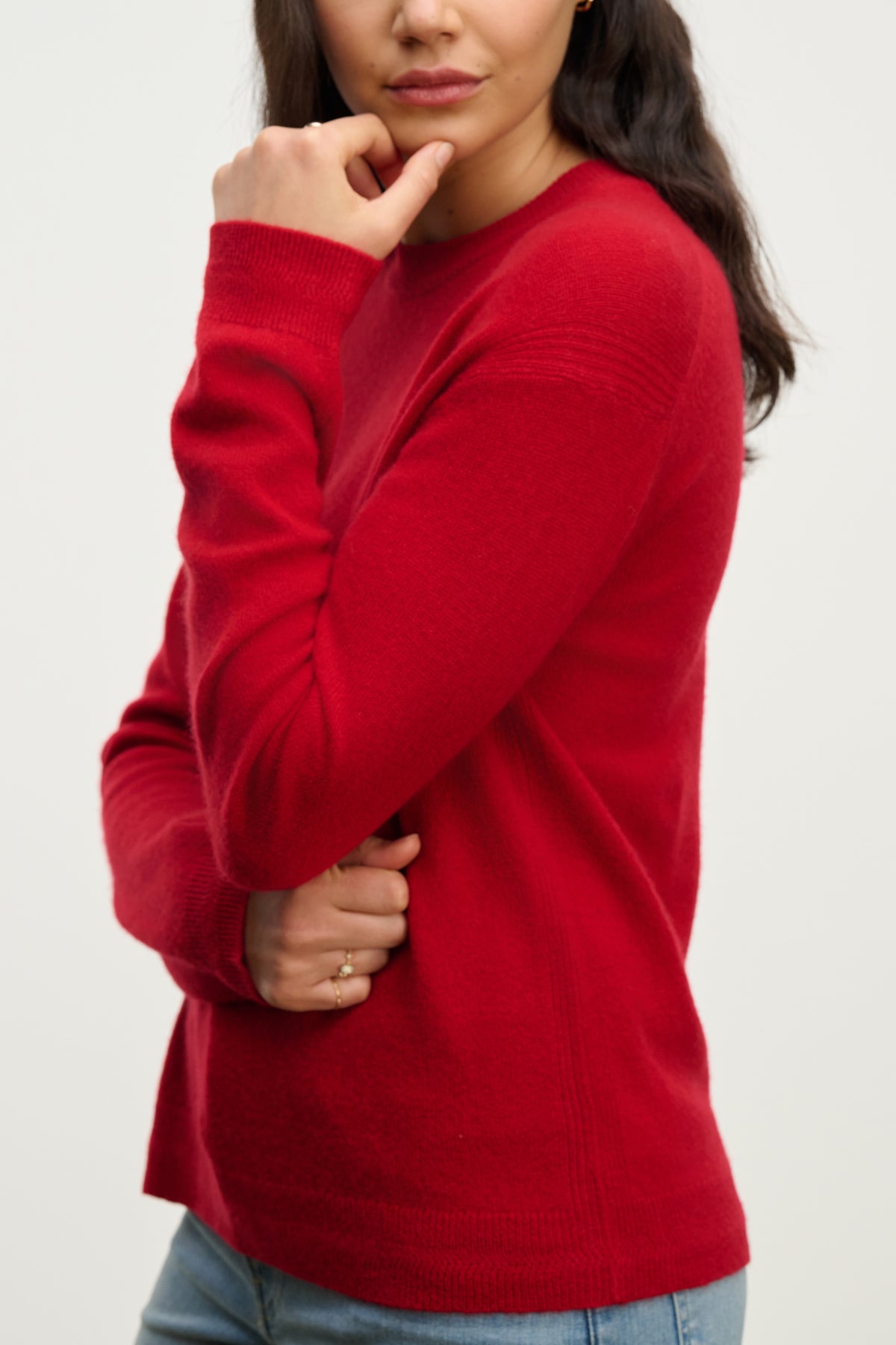   A person with long dark hair wears the CRYSTAL CASHMERE SWEATER by Velvet by Graham & Spencer in red, featuring ribbed trims, paired with blue jeans. They stand arms crossed, facing slightly to the side against a plain background. 