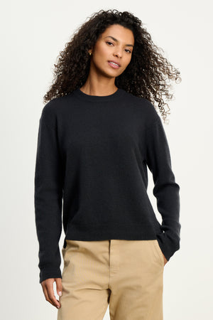 A woman with curly hair wears the Velvet by Graham & Spencer CRYSTAL CASHMERE SWEATER—black with ribbed trims and a crew neckline—paired with beige pants, standing against a plain light background.