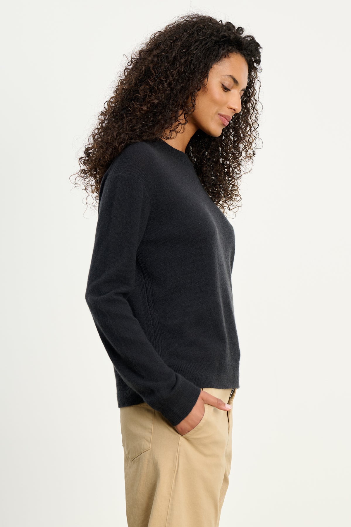 A woman with curly hair stands in profile, wearing Velvet by Graham & Spencer's CRYSTAL CASHMERE SWEATER in black with ribbed trims and beige pants, one hand in her pocket against a plain white background.-39815197163713