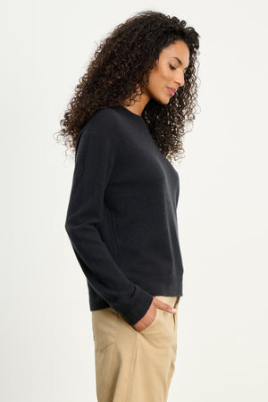 A woman with curly hair stands in profile, wearing Velvet by Graham & Spencer's CRYSTAL CASHMERE SWEATER in black with ribbed trims and beige pants, one hand in her pocket against a plain white background.