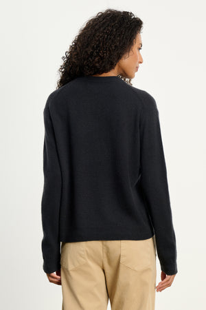 A person with curly hair is seen from the back wearing the CRYSTAL CASHMERE SWEATER by Velvet by Graham & Spencer in black, featuring ribbed trims, paired with beige pants, and standing against a plain light background.