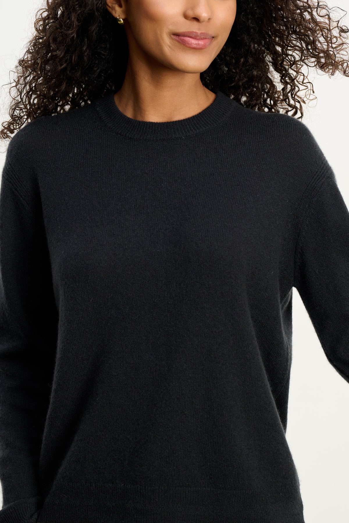 A person with curly hair models the Velvet by Graham & Spencer CRYSTAL CASHMERE SWEATER—a plain black cashmere piece featuring ribbed trims and a classic crew neckline—shown from shoulders to waist against a neutral background.-39815197065409