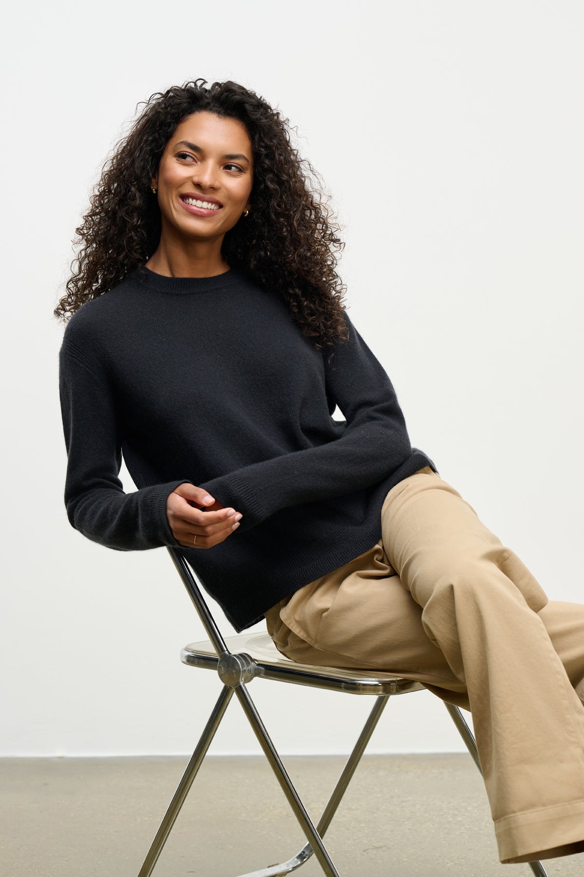 A woman with curly hair, wearing Velvet by Graham & Spencer’s CRYSTAL CASHMERE SWEATER in black and beige pants, smiles while seated on a metal folding chair against a plain light background.-39815197032641