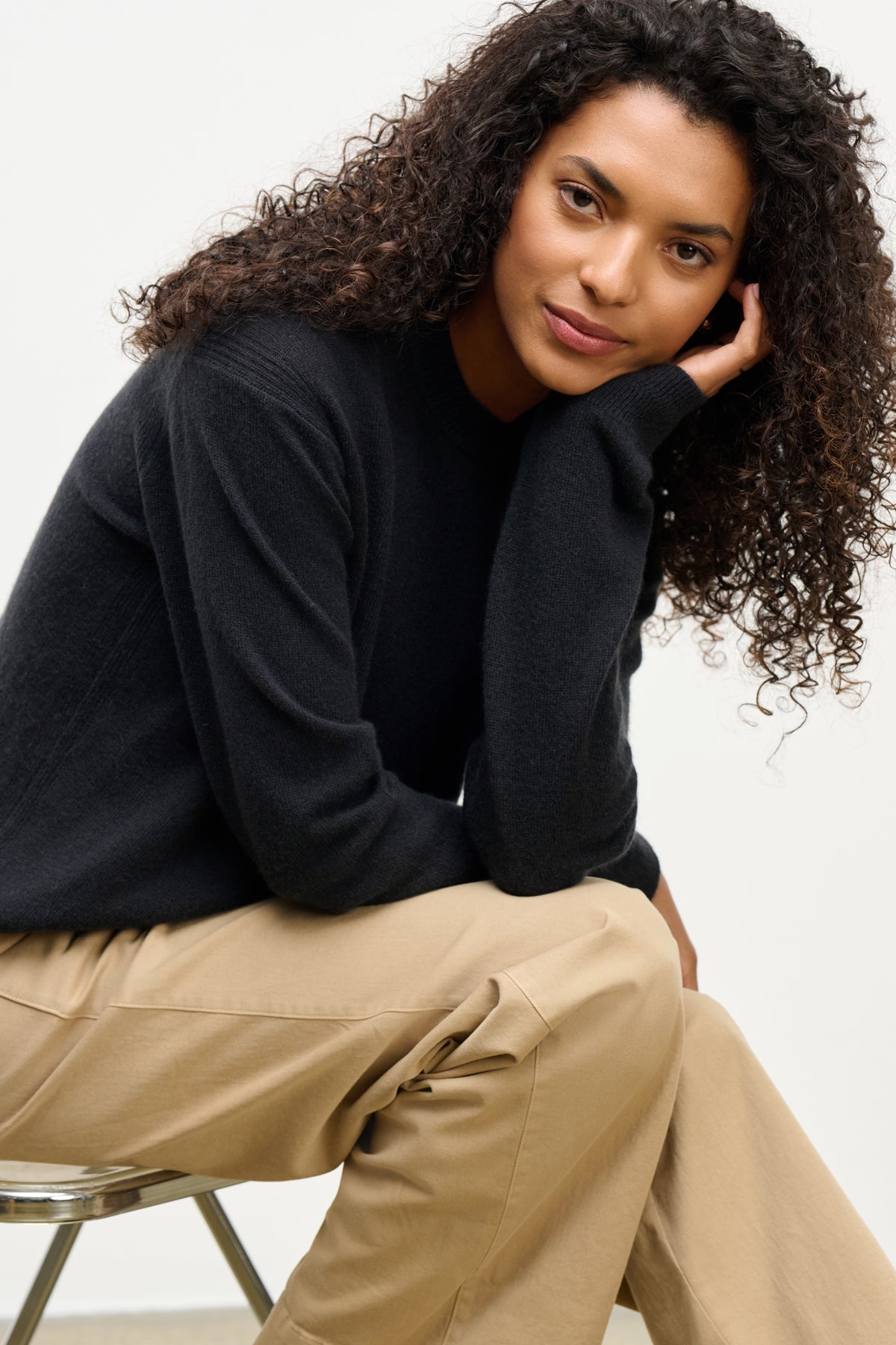 A woman with curly hair wears the Velvet by Graham & Spencer CRYSTAL CASHMERE SWEATER in black with a crew neckline and beige pants, sitting on a stool and resting her head on her hand while looking at the camera.-39815196967105
