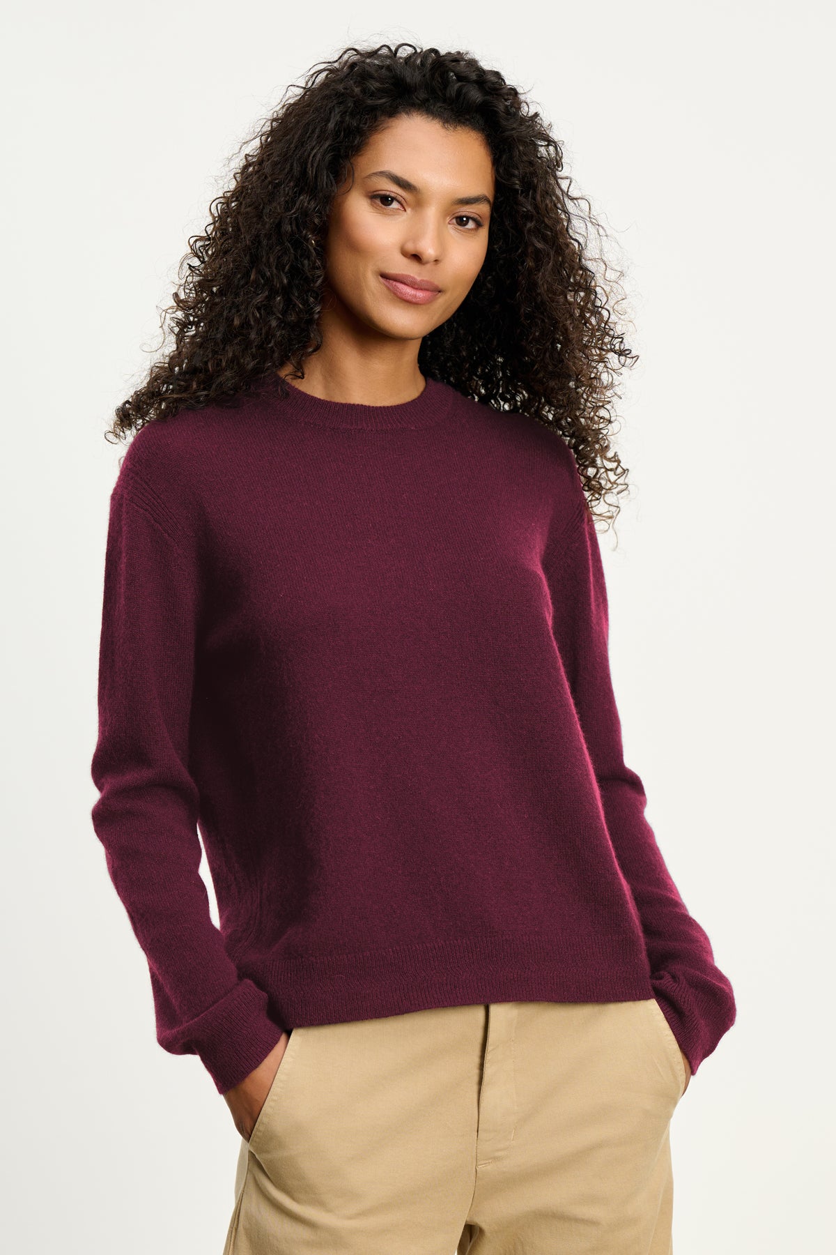 A woman with curly hair wears the Velvet by Graham & Spencer CRYSTAL CASHMERE SWEATER in maroon, featuring a crew neckline and ribbed trims, styled with beige pants and her hands in her pockets against a plain light background.-39838379245761