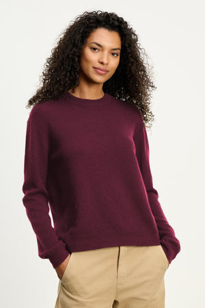 A woman with curly hair wears the Velvet by Graham & Spencer CRYSTAL CASHMERE SWEATER in maroon, featuring a crew neckline and ribbed trims, styled with beige pants and her hands in her pockets against a plain light background.