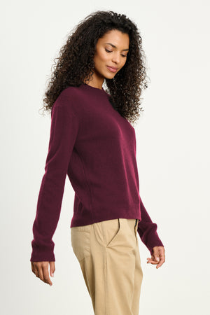 A woman with curly hair wears the Velvet by Graham & Spencer CRYSTAL CASHMERE SWEATER in burgundy, featuring long sleeves and ribbed trims, paired with beige pants against a plain light background.