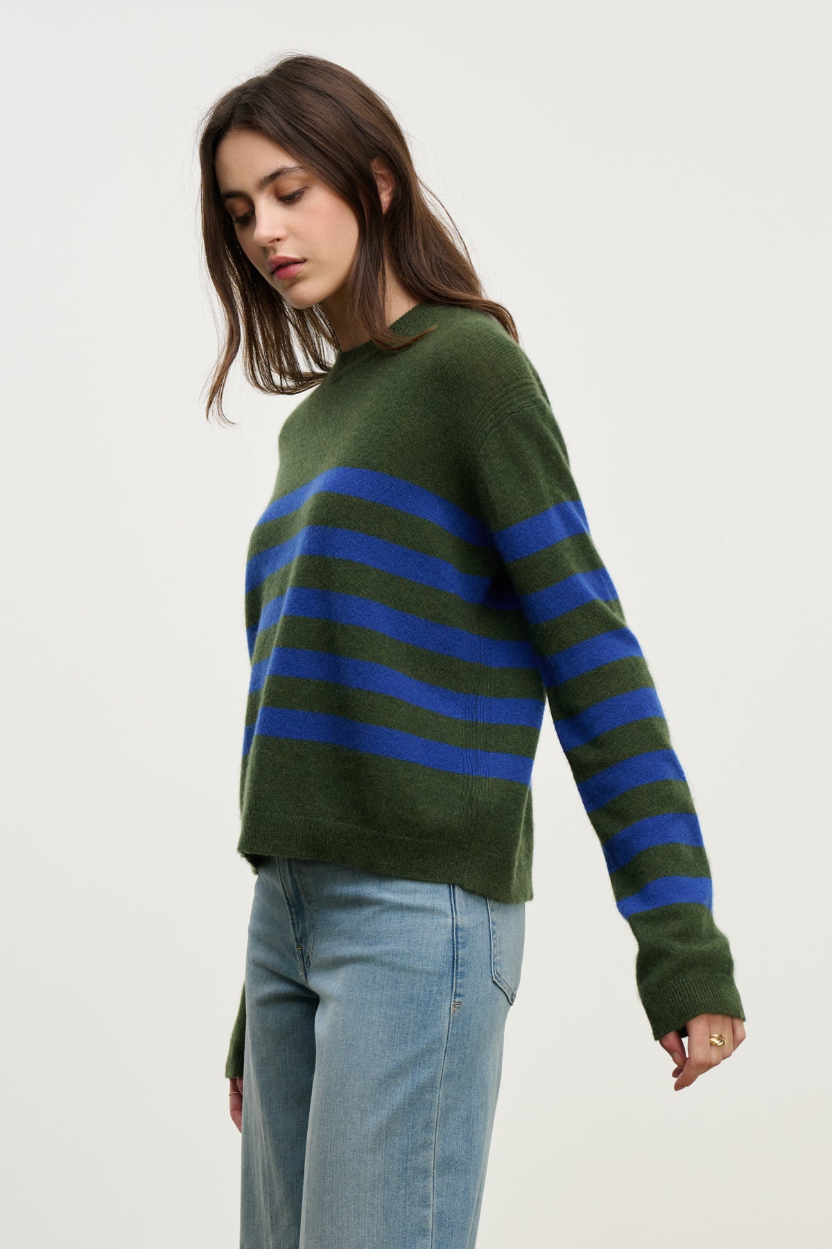 A woman wears the DIANA CASHMERE SWEATER by Velvet by Graham & Spencer in green and blue stripes, paired with light blue jeans, standing against a plain light background.-39975057195201