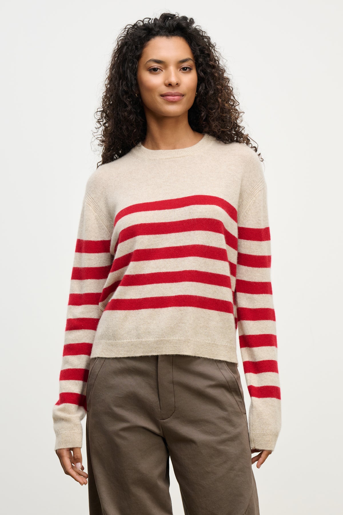 A woman wears the Velvet by Graham & Spencer DIANA CASHMERE SWEATER—beige with red stripes—in a relaxed fit, paired with brown pants, against a plain light background.-39975057129665