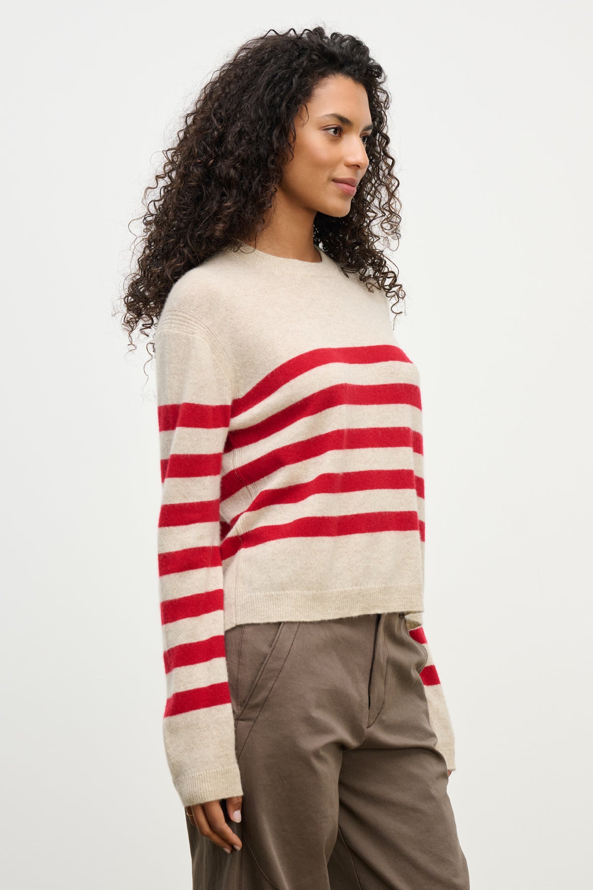 A person wears the DIANA CASHMERE SWEATER by Velvet by Graham & Spencer—a relaxed-fit beige sweater with red stripes—paired with brown pants, standing against a plain light background.-39975057096897