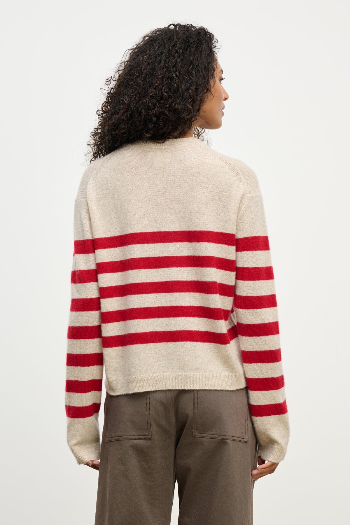 A person with curly hair is seen from the back wearing the DIANA CASHMERE SWEATER by Velvet by Graham & Spencer—a relaxed fit beige sweater with horizontal red stripes—paired with brown pants.-39975057064129
