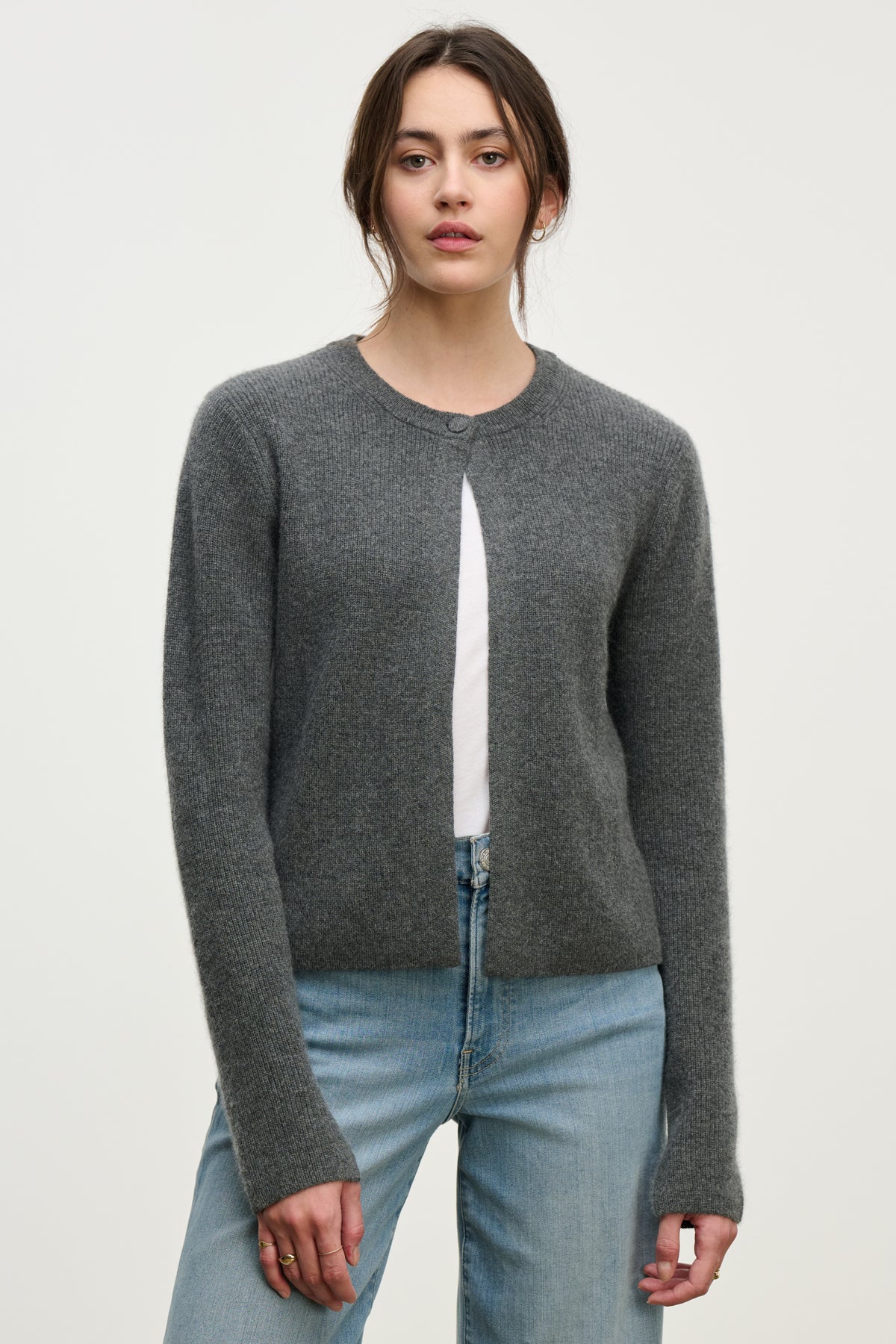 A person wears the TINA CASHMERE CARDIGAN by Velvet by Graham & Spencer—a gray, ribbed, long-sleeved cardigan—over a white shirt and blue jeans, standing against a plain light background.-39814940623041