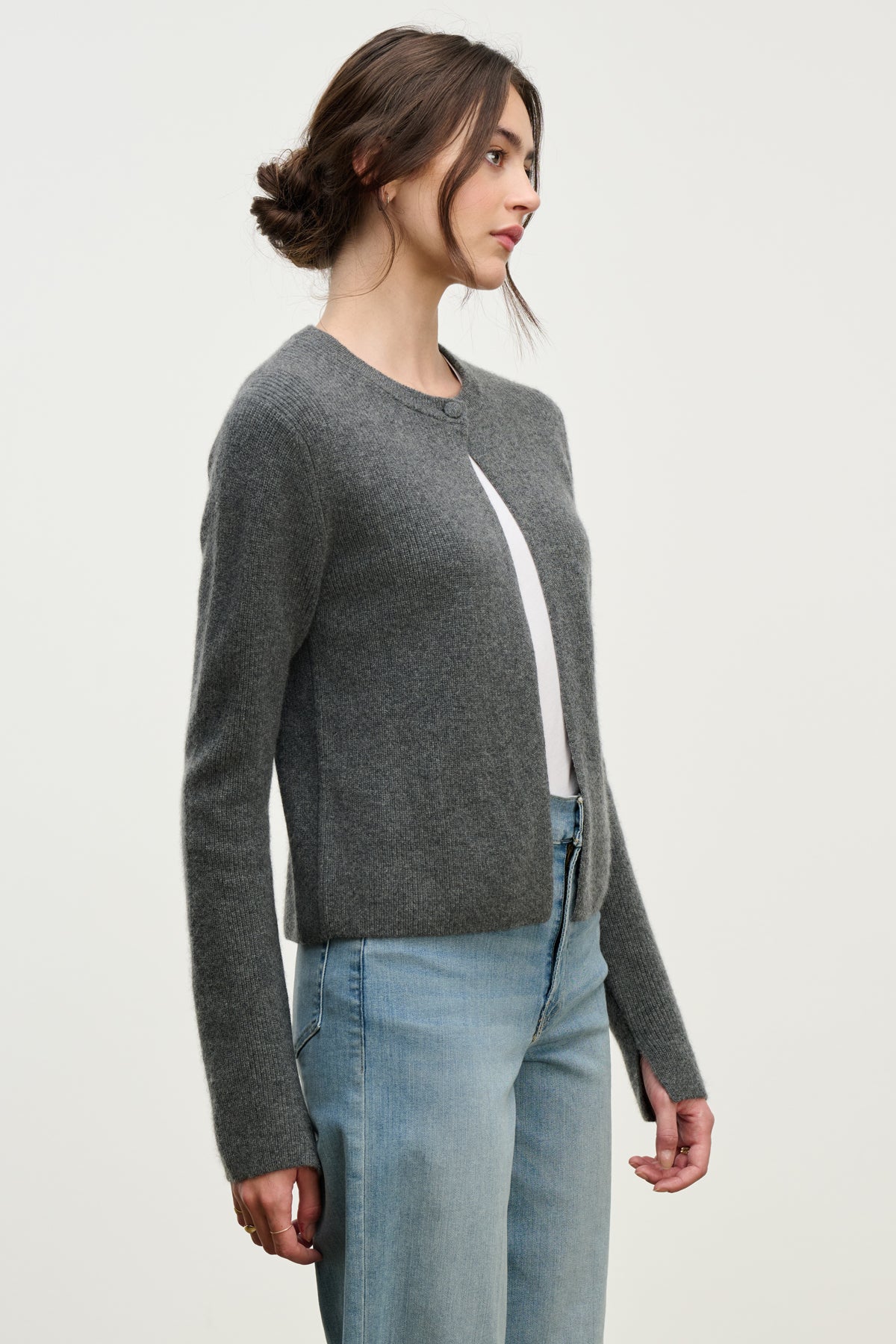 A woman stands in profile against a white background, wearing the Velvet by Graham & Spencer TINA CASHMERE CARDIGAN in gray over a white top and light blue jeans.-39814940524737