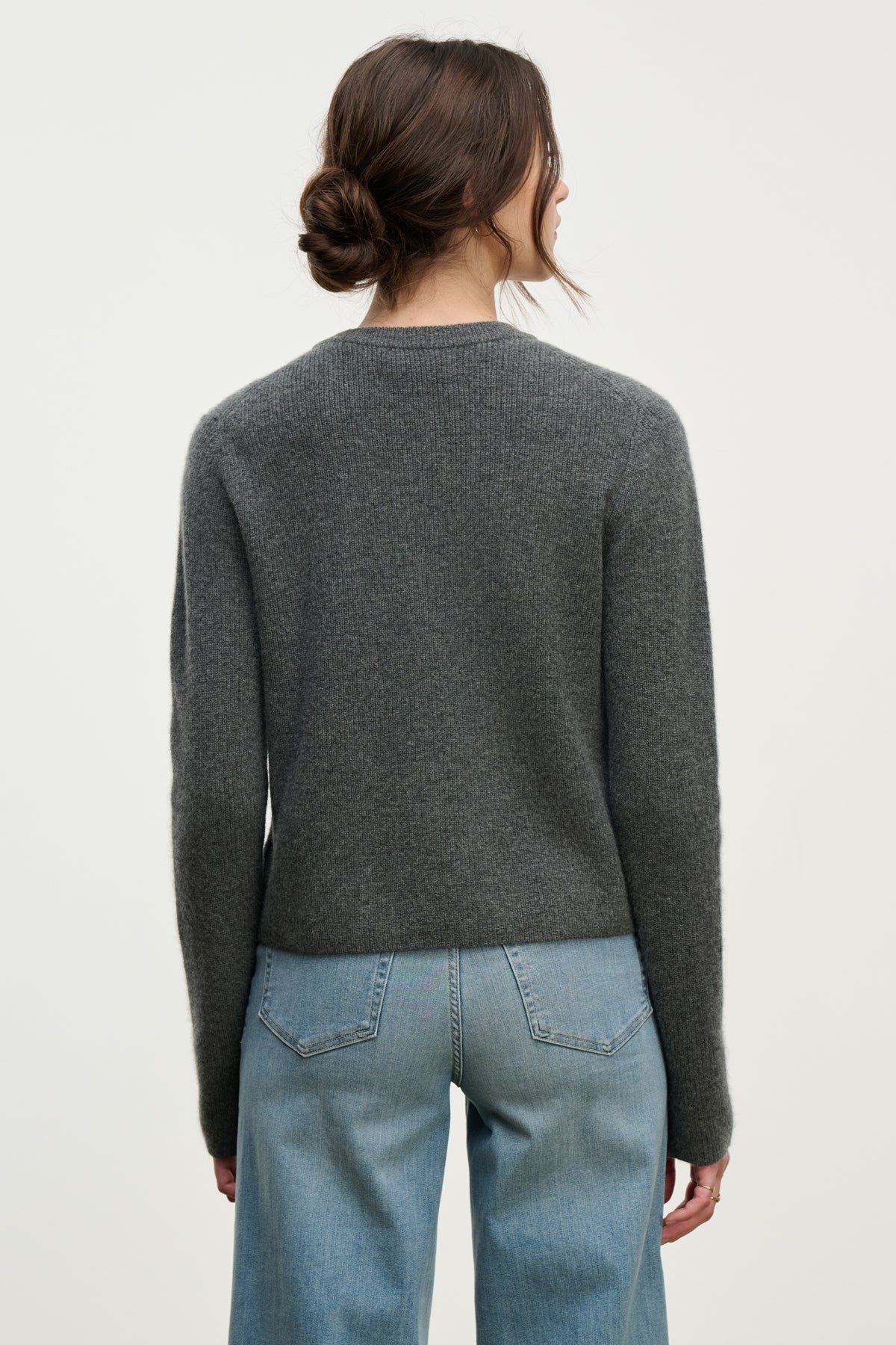 A woman with brown hair in a low bun wears the TINA CASHMERE CARDIGAN by Velvet by Graham & Spencer with light blue jeans, standing with her back to the camera—a versatile piece for effortless layering.-39814940491969