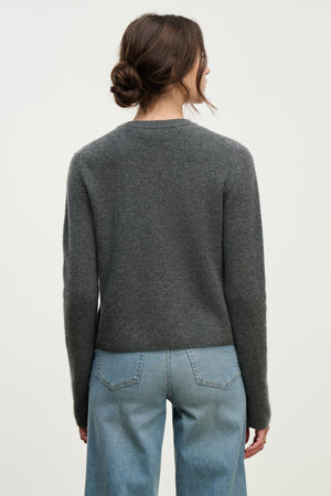 A woman with brown hair in a low bun wears the TINA CASHMERE CARDIGAN by Velvet by Graham & Spencer with light blue jeans, standing with her back to the camera—a versatile piece for effortless layering.