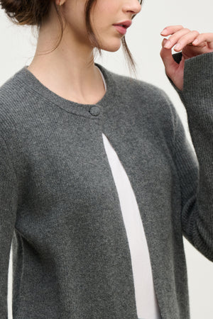 A woman wearing the TINA CASHMERE CARDIGAN by Velvet by Graham & Spencer—a grey, long-sleeve, buttoned cardigan with a subtle ribbed texture over a white top—makes it a perfect layering piece for any wardrobe.