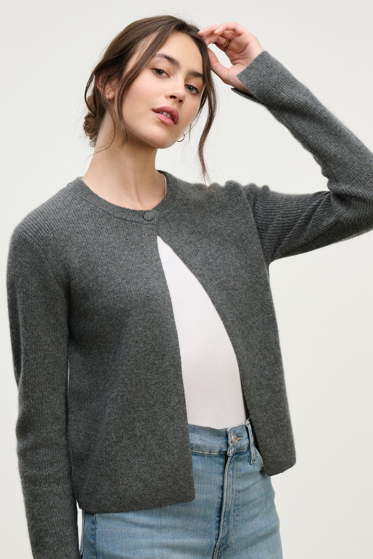 A woman wears the TINA CASHMERE CARDIGAN by Velvet by Graham & Spencer in grey over a white top with light blue jeans, standing against a plain background and raising one hand to her head.-39814940393665