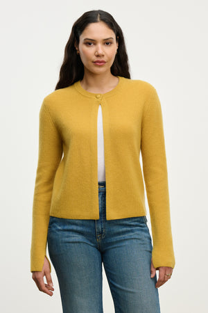 A woman models the TINA CASHMERE CARDIGAN by Velvet by Graham & Spencer in mustard yellow over a white top and blue jeans, standing against a plain background to showcase this perfect layering piece.