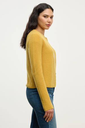 A woman with long dark hair wears the TINA CASHMERE CARDIGAN by Velvet by Graham & Spencer in yellow with blue jeans, standing sideways against a plain light background.