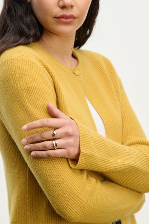 A woman in the Velvet by Graham & Spencer TINA CASHMERE CARDIGAN, featuring a ribbed mustard yellow knit, stands with her arms crossed, showcasing two gold rings—a perfect layering piece for any outfit.