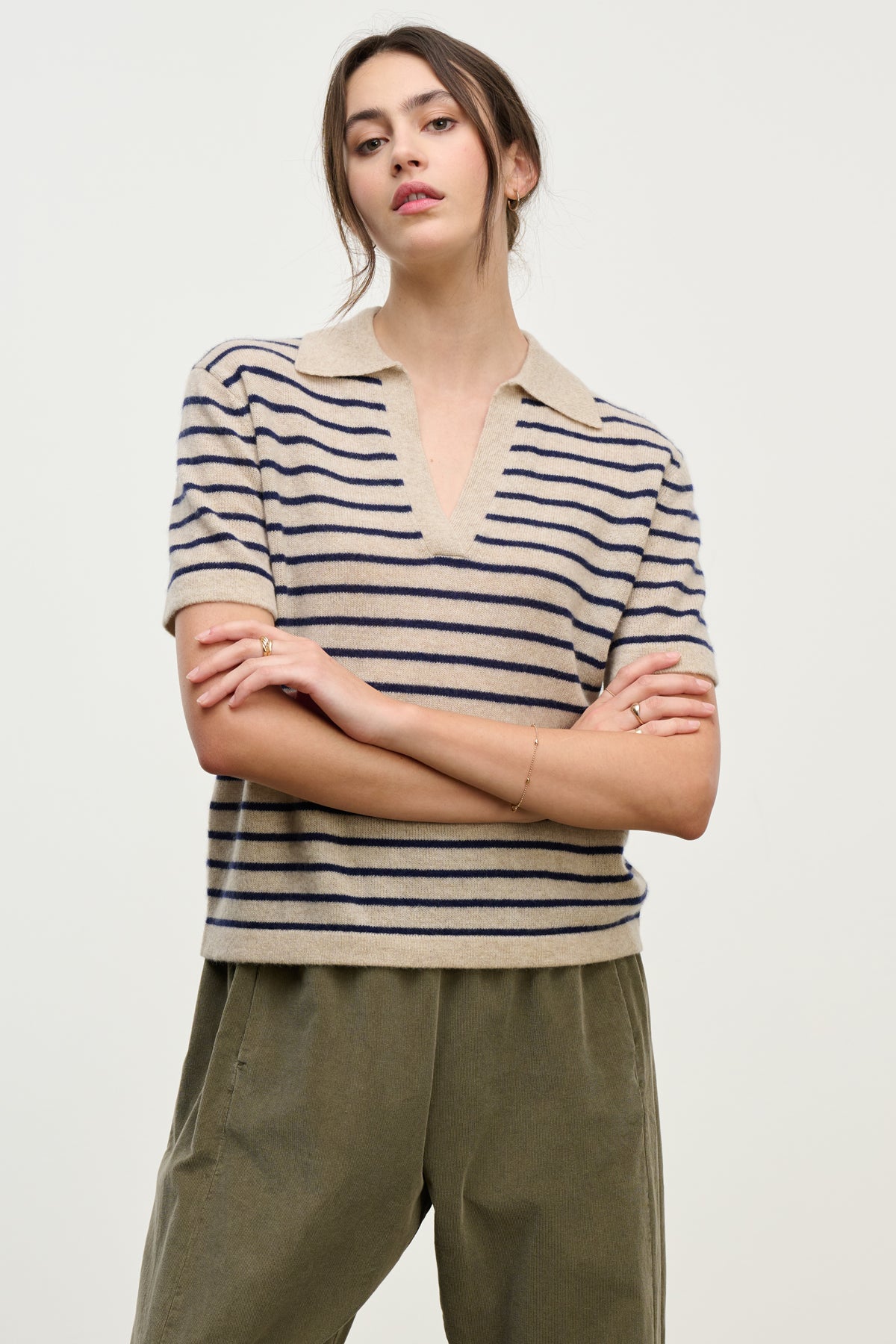   A woman stands with arms crossed against a plain background, wearing the YAEL CASHMERE SWEATER POLO by Velvet by Graham & Spencer in beige and navy stripes, paired with olive green pants. 