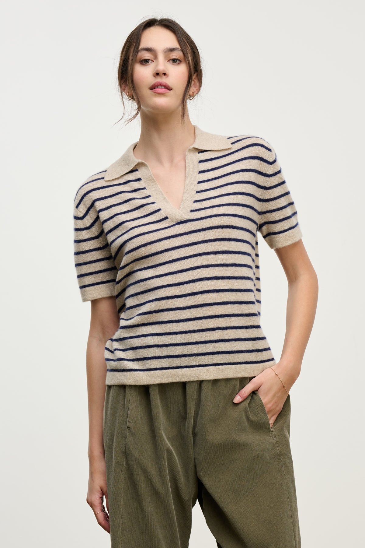   A woman wears the YAEL CASHMERE SWEATER POLO by Velvet by Graham & Spencer—a cropped beige cashmere sweater with navy stripes and a collar—paired with loose olive green pants, standing against a plain light background. 