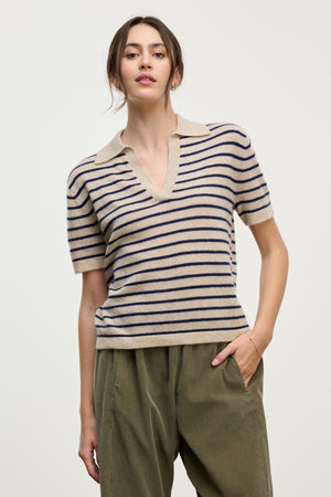 A woman wears the YAEL CASHMERE SWEATER POLO by Velvet by Graham & Spencer—a cropped beige cashmere sweater with navy stripes and a collar—paired with loose olive green pants, standing against a plain light background.